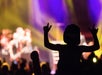 A child wearing a cap is silhouetted against colorful stage lights, raising both hands in a rock and roll gesture at a concert.