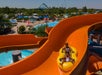 Two people ride a yellow inflatable raft down an orange water slide at a water park, with pools, lounge chairs, and greenery visible in the background.