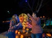 Two people pose playfully near a glowing dinosaur-shaped light display surrounded by pumpkins at night, with festive lights in the background.