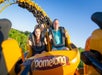 Three people ride a yellow roller coaster called Boomerang, smiling and raising their hands, with green trees and blue sky in the background.