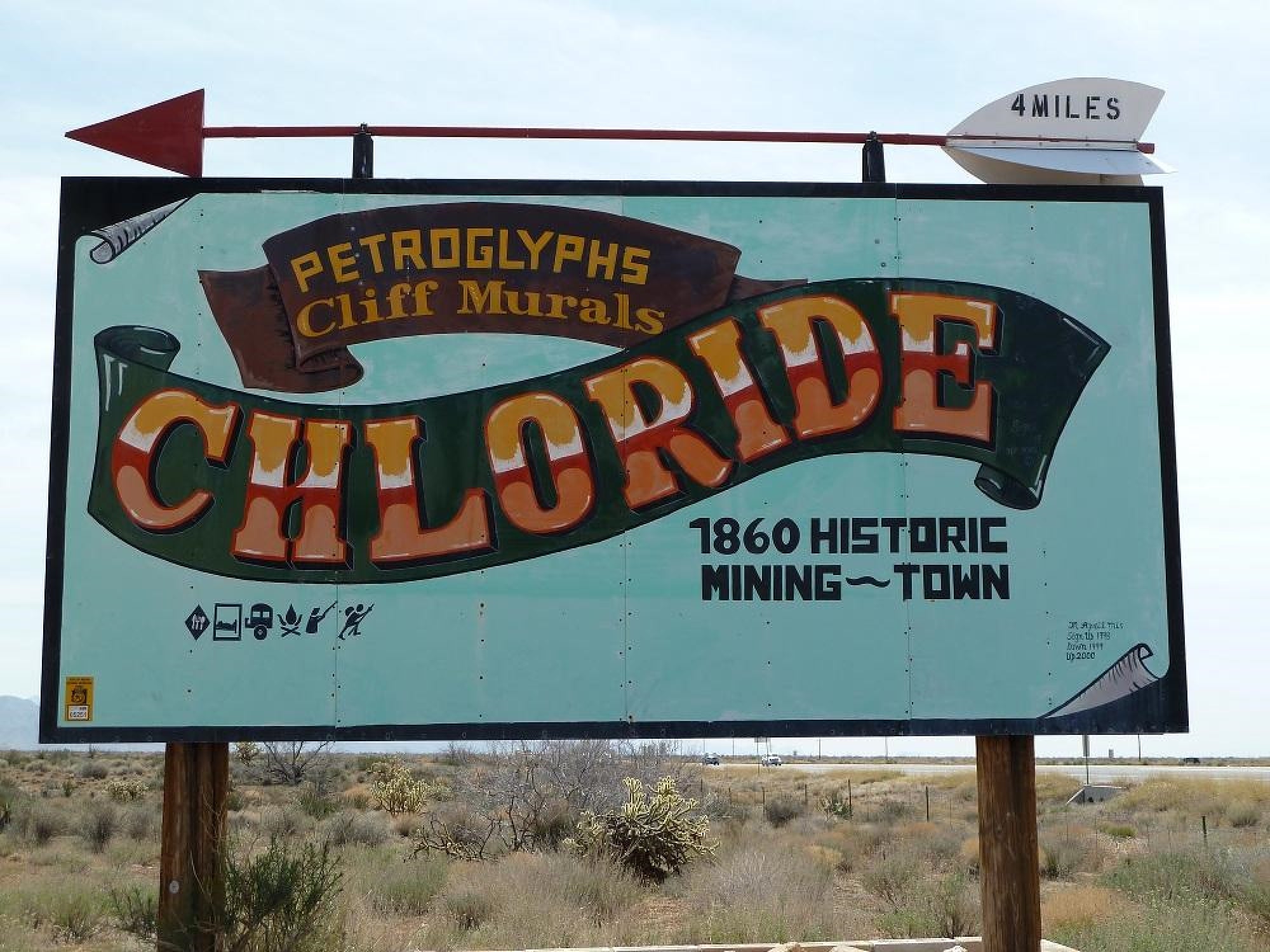 A roadside sign reads “Chloride: 1860 Historic Mining Town” with directions to petroglyphs and cliff murals, and an arrow indicating the town is 4 miles away. Desert landscape in background.