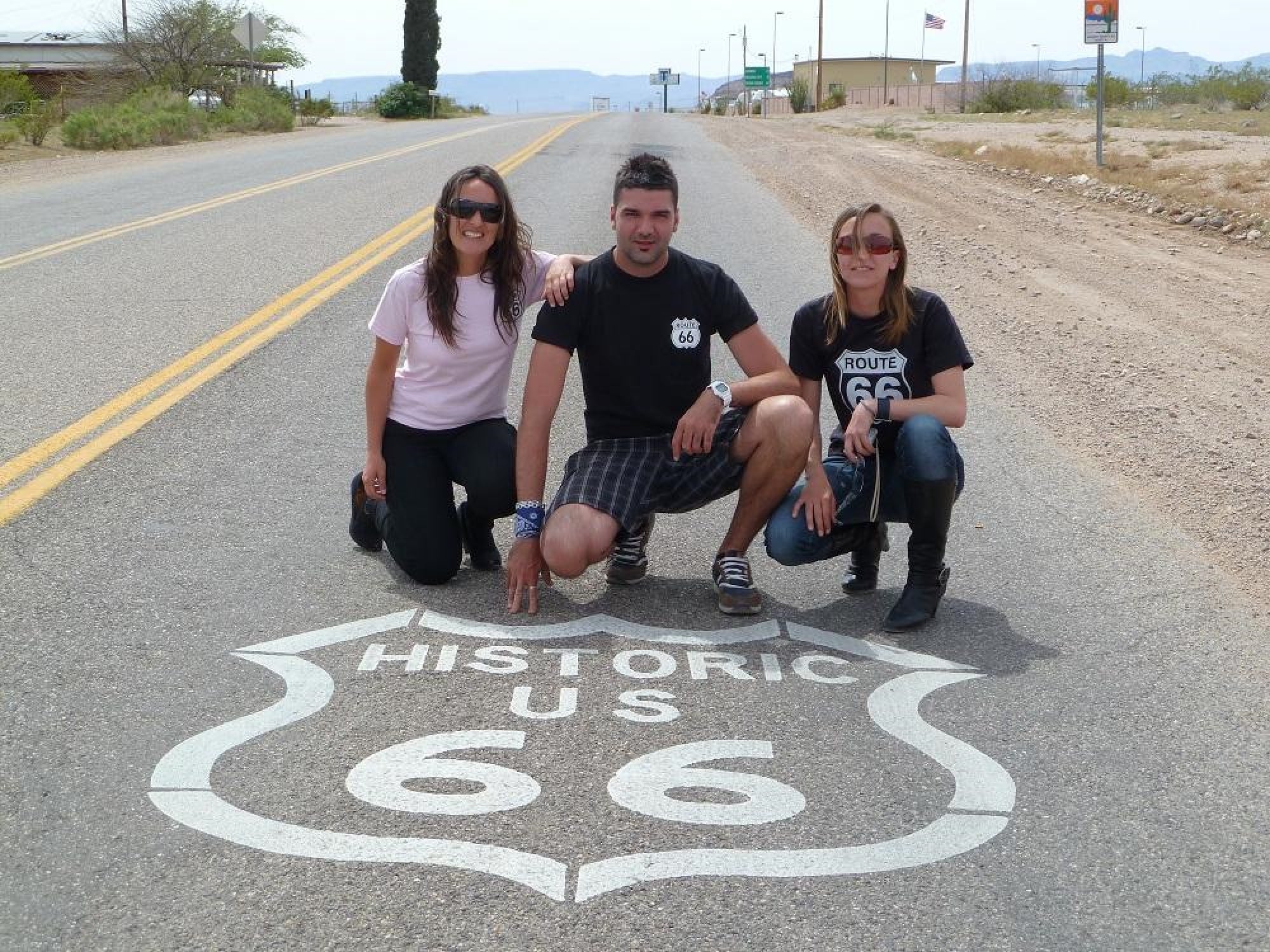 Three people kneel on the pavement next to a "Historic US 66" road sign painted on the street, with a desert landscape and road signs in the background.
