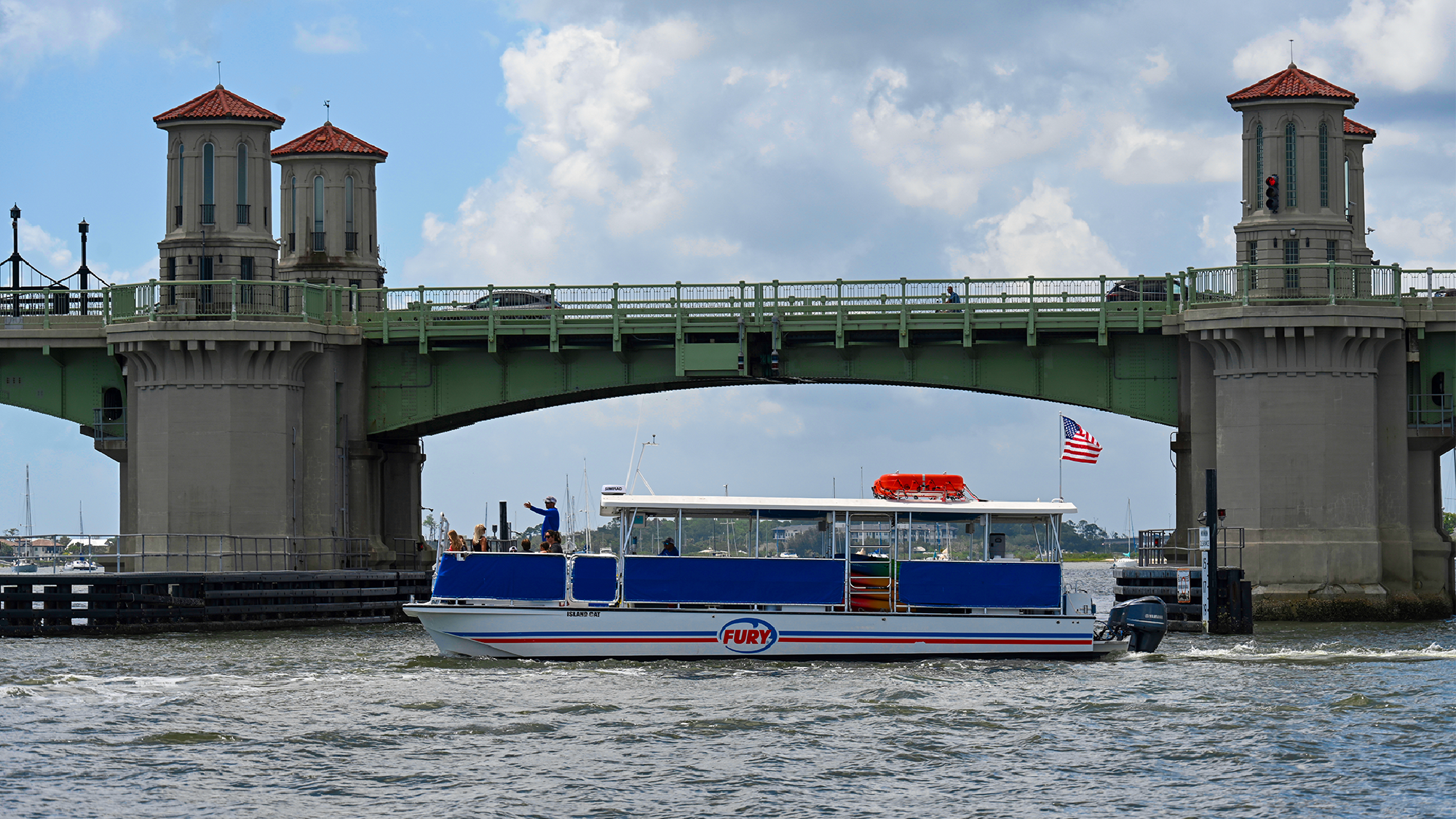 A sightseeing boat labeled "Fury" passes under a green drawbridge with two towers, as people stand on the deck; a U.S. flag flies at the stern.
