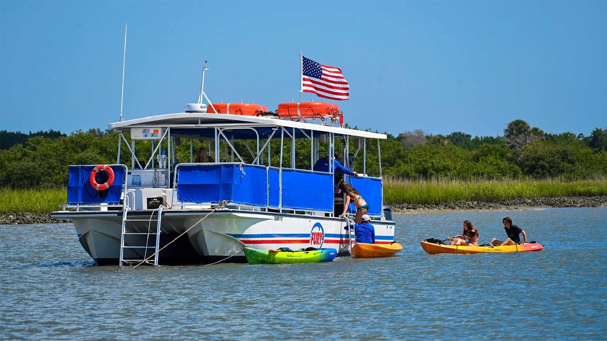 A large boat with an American flag is anchored near the shore as two people in kayaks approach, with green trees and blue sky in the background.