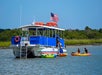 A large boat with an American flag is anchored near the shore as two people in kayaks approach, with green trees and blue sky in the background.