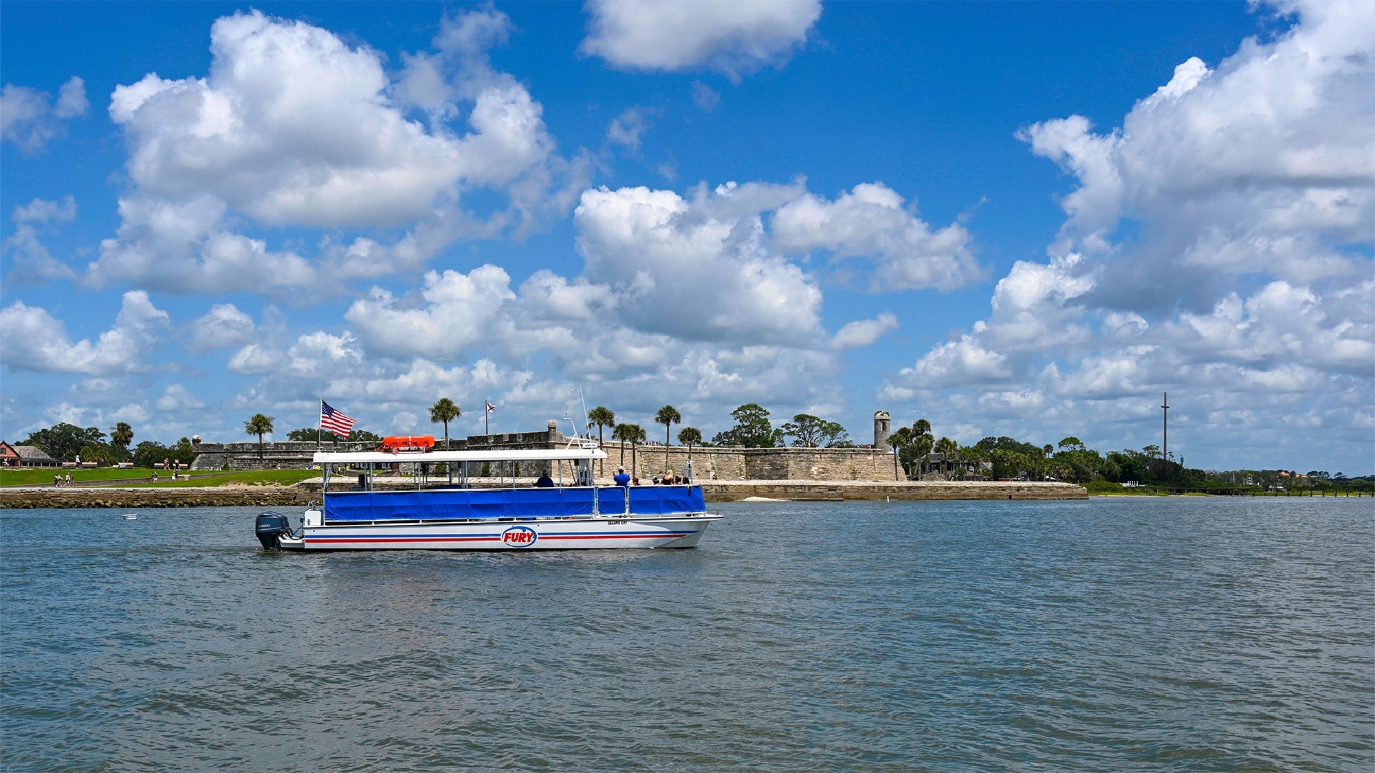 A blue and white tour boat with an American flag sails on the water near a historic stone fort with palm trees under a partly cloudy sky.