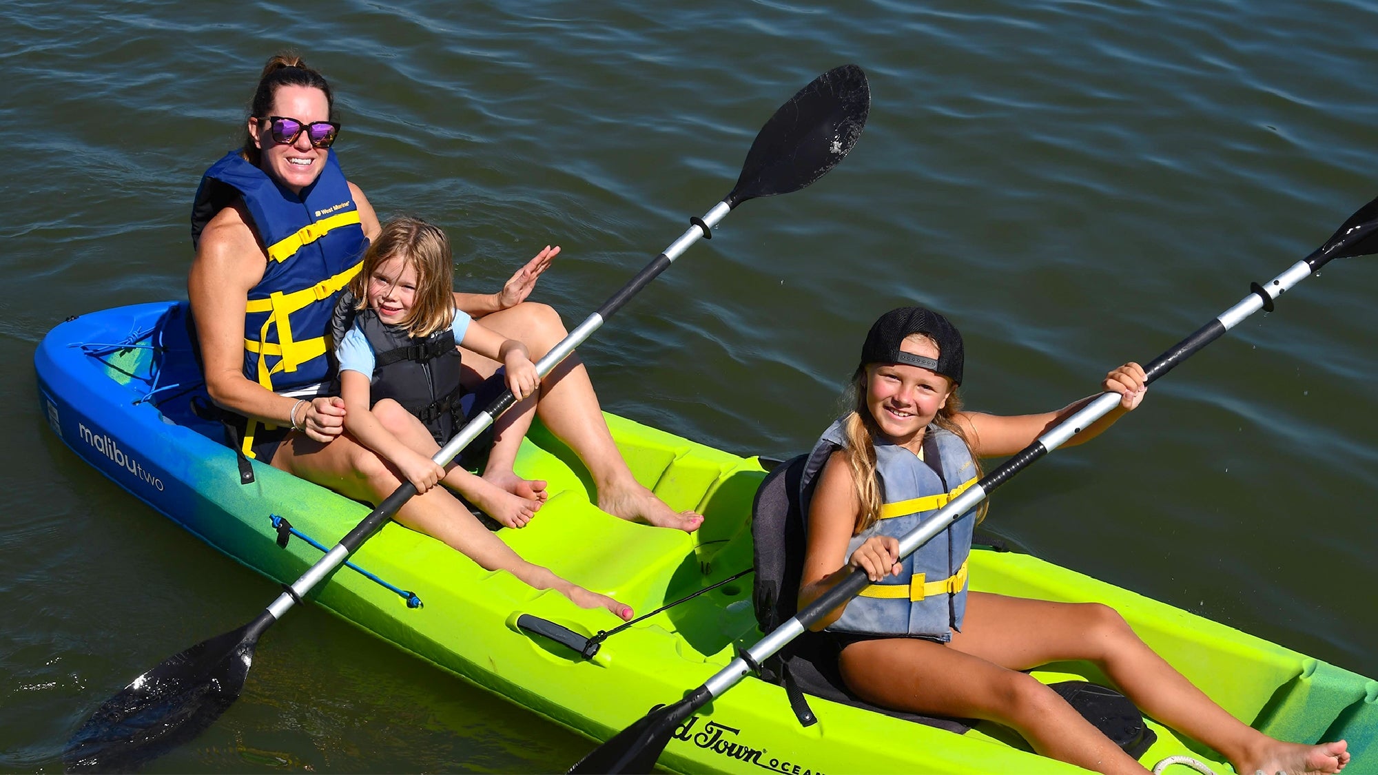 Two girls and an adult wearing life jackets paddle a green kayak together on calm water under bright sunlight.