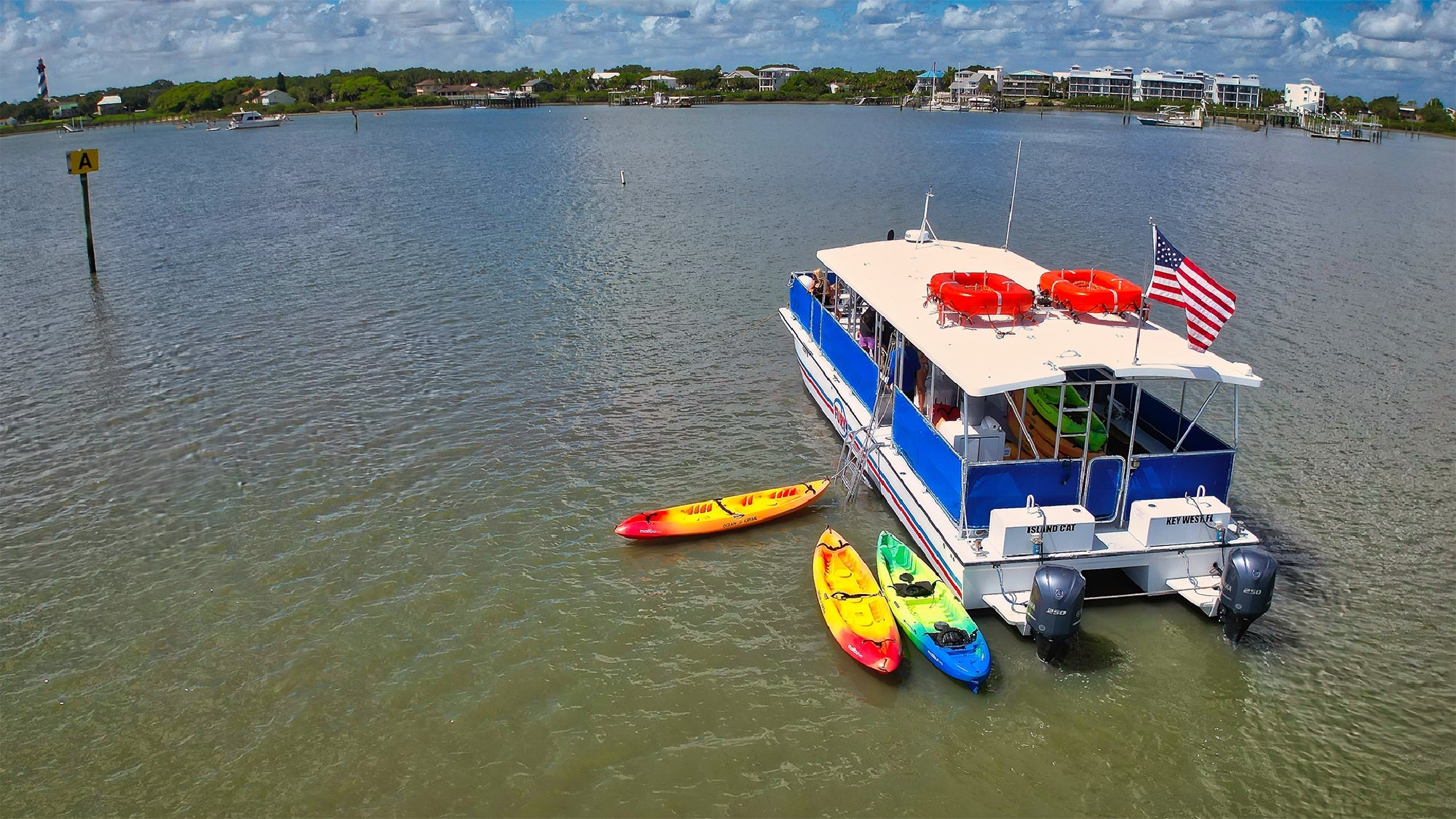 A blue and white boat with an American flag is anchored on calm water, with three colorful kayaks beside it; shoreline and buildings are visible in the background.