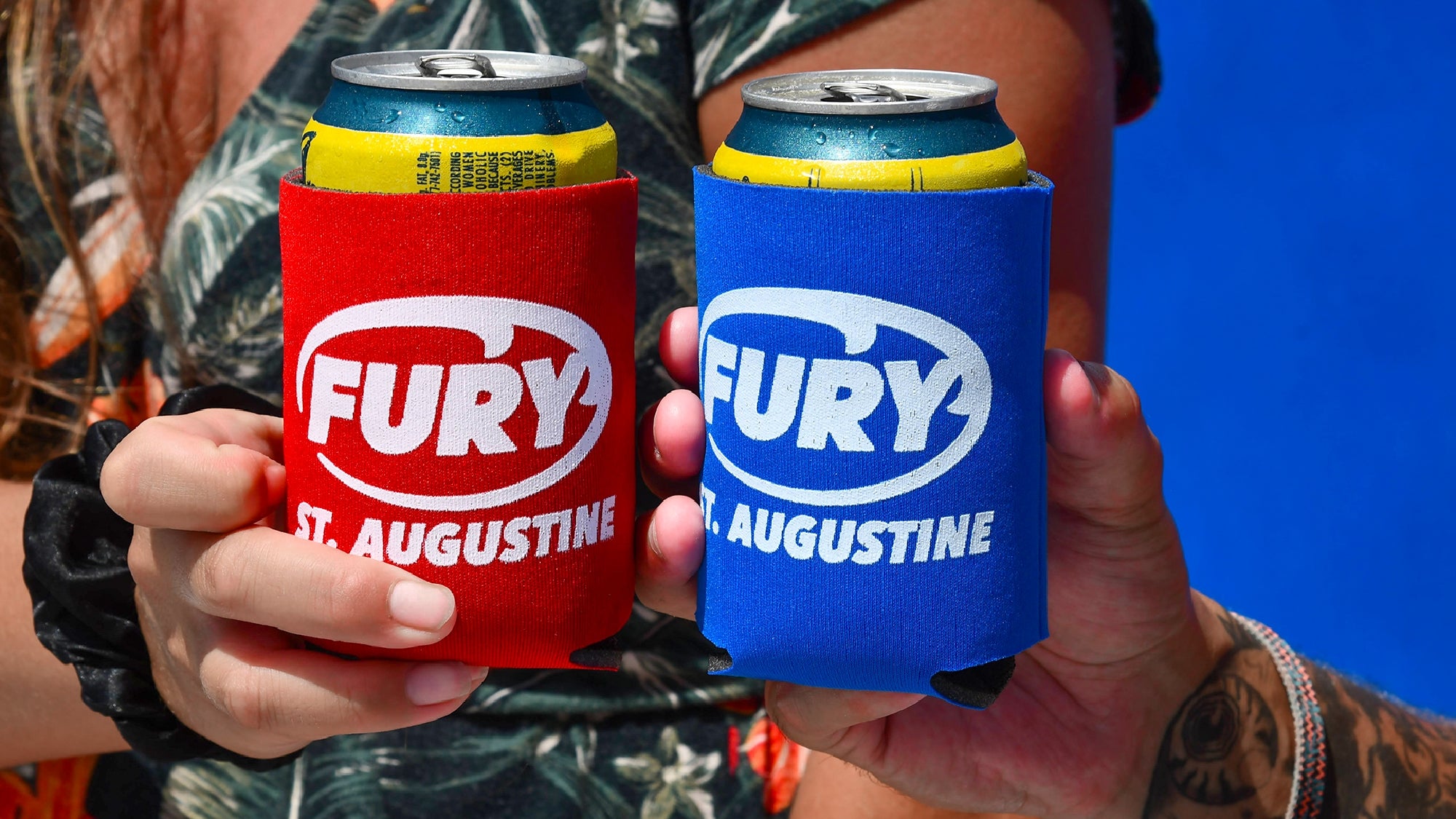 Two people hold canned drinks in red and blue "FURY ST. AUGUSTINE" can coolers, with a blue background and part of a floral shirt visible.