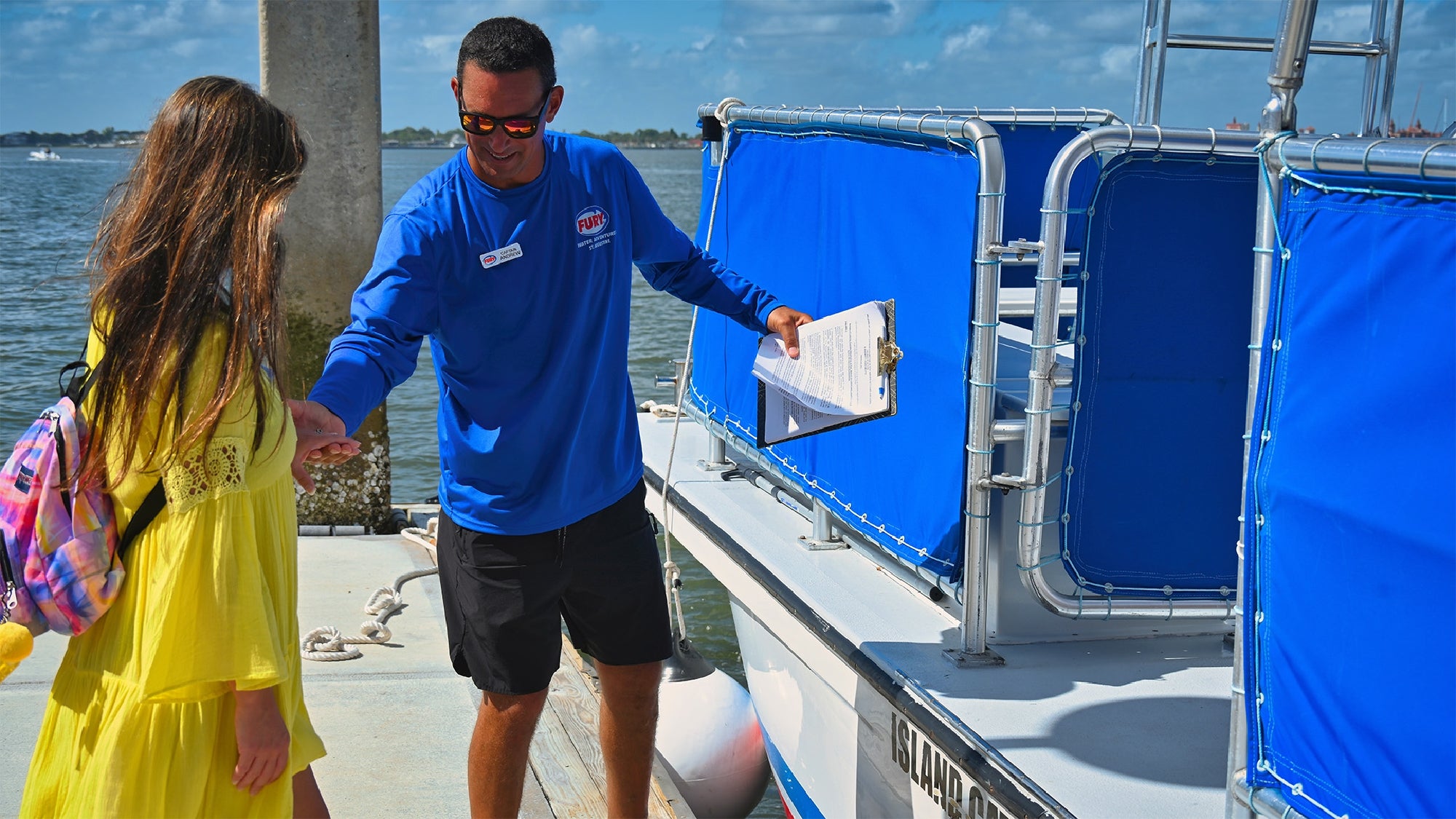 A man in a blue shirt holds a clipboard and gestures as he assists a woman in a yellow dress onto a docked boat.