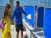 A man in a blue shirt holds a clipboard and gestures as he assists a woman in a yellow dress onto a docked boat.