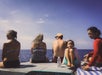 Five people sit on a dock or boat facing the ocean, with a partly cloudy sky in the background. A water bottle and towel are visible in the foreground.