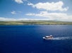 A small boat moves across deep blue water near a green, low-lying coastline under a partly cloudy sky.