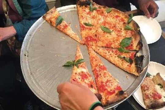 A large cheese pizza with basil leaves on a metal tray, several slices missing, and people taking slices with their hands.
