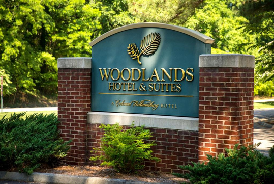 Sign for Woodlands Hotel & Suites, a Colonial Williamsburg Hotel, set in a landscaped area with greenery in the background.
