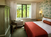 A hotel room with a double bed covered in white and red linens, a window with curtains, an armchair, an armoire, and a wall-mounted lamp. The walls have floral wallpaper.