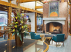Cozy lounge with brick fireplace, two teal chairs, and a large floral arrangement on a table. Warm lighting and contemporary decor.