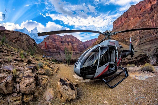 A helicopter is parked on rocky terrain in a canyon landscape under a partly cloudy sky.