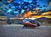 A silver, black and red Maverick Airbus helicopter on the ground with and bright blue and orange cloudy sky overhead.