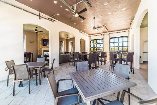 Covered outdoor patio with several tables and chairs, ceiling fans, and large windows letting in natural light. The space appears clean and well-lit.