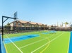 Outdoor multi-use sports court with basketball hoops and tennis net, surrounded by fencing, with residential buildings and palm trees in the background under a clear blue sky.