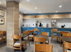 Hotel breakfast area with wooden chairs and tables. Blue counter has coffee machines, toaster, and a selection of breakfast items against a white tiled wall. Ceiling lights are on.