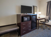 Hotel room with a TV on a cabinet, a microwave, a mini-fridge, a desk with a lamp, and a modern chair. Neutral-colored walls and carpet.