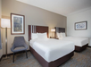 Hotel room with two double beds, modern decor, a floor lamp, and artwork on the walls. A gray upholstered chair is placed next to the beds.