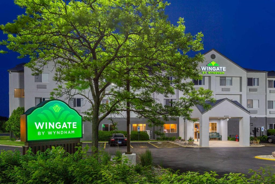 Exterior of a Wingate by Wyndham hotel at night, with illuminated signage and a tree in the foreground.
