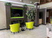 Lobby area with a wall-mounted TV, two lime green chairs, a small black table, and a large potted plant. Neutral tones decorate the space.
