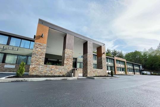 A modern building with a stone-accented facade and large windows, set against a clear sky. The foreground shows a wide, empty asphalt driveway.