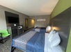 Hotel room with two double beds, a TV, a microwave, a mini-fridge, and a green chair. Walls are gray with a green accent, and artwork hangs above the beds.