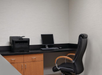 Office corner with a black chair, wooden desk, computer monitor, keyboard, and printer set against a light gray wall.