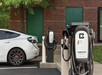 Electric vehicle charging station with two chargers, one connected to a white car. Brick building and green doors in the background.
