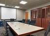 A conference room with a large rectangular table, surrounded by black chairs. A projector screen is on the wall, and notepads with pens are placed at each seating spot.