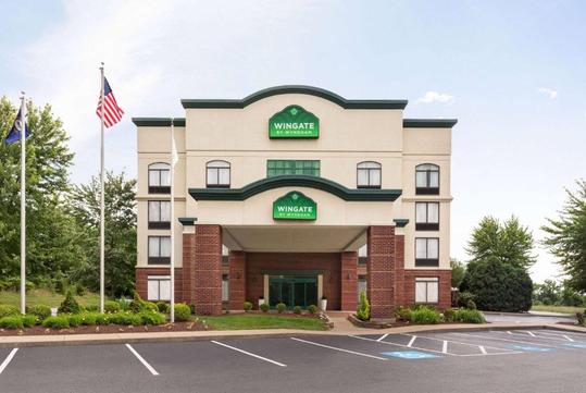 Front view of a four-story Wingate hotel with American and state flags, surrounded by trees and parking spaces.