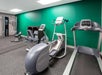 Small gym with exercise machines including a treadmill, recumbent bike, and elliptical trainer on a rubber mat floor, against a green wall.