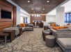 Modern lounge with patterned carpet, gray seating, and orange cushions. Wall-mounted TV on the left, dining table with chairs in the back, and ceiling lights above.