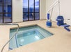 An indoor hot tub with a blue and white accessibility lift on the side, large windows in the background, and a metal handrail on the left.
