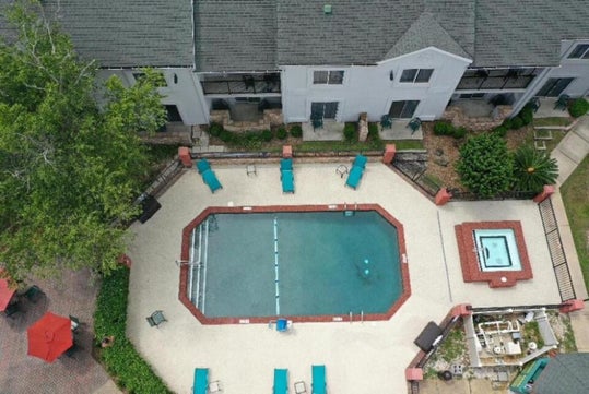 Aerial view of a rectangular outdoor pool with lounge chairs, a hot tub, and surrounding patio area, adjacent to a multi-unit residential building.