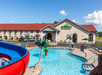 Outdoor swimming pool with water slide at a hotel, featuring palm tree decorations. Hotel building in the background, clear blue sky above.