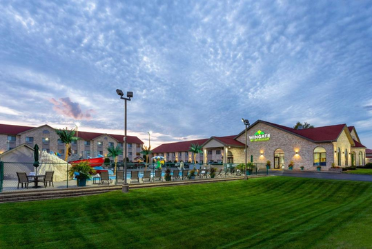 Hotel building with red roofs, outdoor pool area, and green lawn under a cloudy sky.