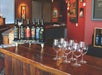 Six wine bottles are displayed on a small rack beside eight empty wine glasses on a wooden counter in a warmly lit tasting room with art on the walls.