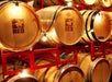 Stacks of wooden wine barrels on red metal racks, with lights strung overhead. One barrel displays the label "Love That Red.