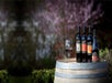 Four bottles of red wine and a filled wine glass are displayed on top of a wine barrel outdoors, with a blurred background of trees and foliage.