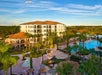 Arial view of a resort with a large pool, palm trees, and a multi-story building under a clear blue sky.