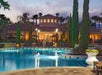 Luxurious poolside with lounge chairs, surrounded by tall trees and a villa-style house in the background, at sunset.