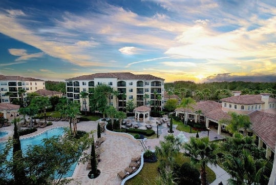 Aerial view of a resort featuring a large swimming pool, surrounded by palm trees and multi-story buildings, under a colorful sky at sunset.