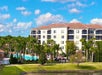 A large white building with balconies is surrounded by palm trees. There is a pool area with lounge chairs in the foreground. The sky is partly cloudy.