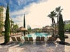 Outdoor pool area with lounge chairs, umbrellas, and tall palm trees under a partly cloudy sky.